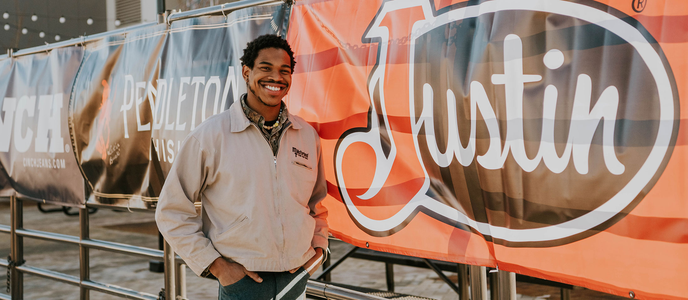 Man stands next to a fence with Justin Boots Banner on the fence.